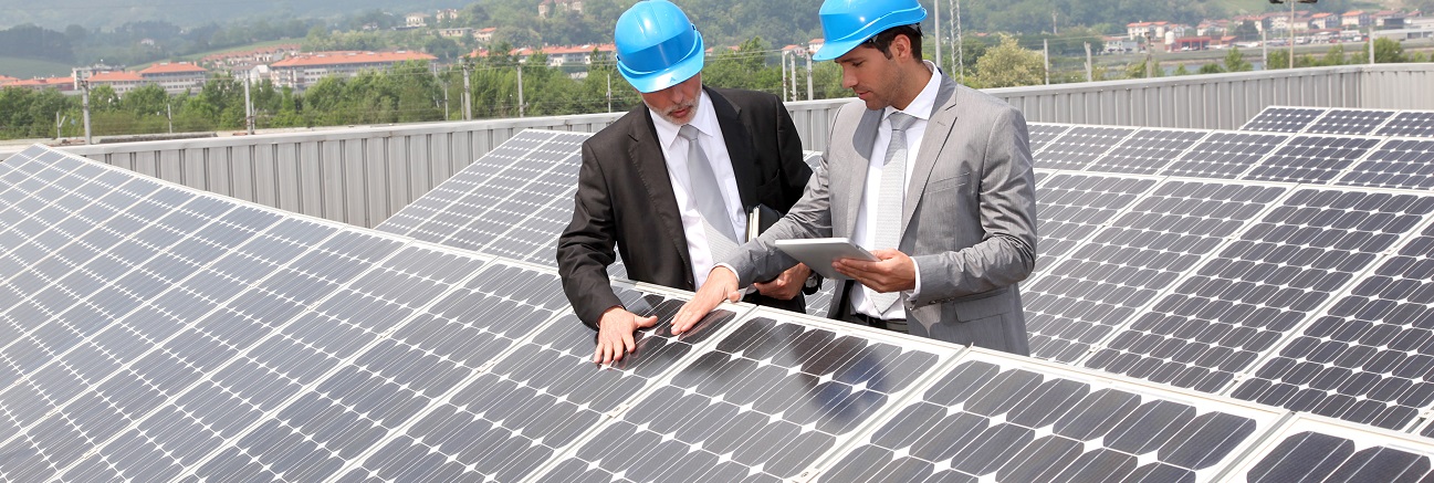 Zwei Techniker auf dem Dach einer Fertigungshalle begutachten die darauf installierten Solarpanele. Der rechts abgebildete Techniker hält ein Tablet in der Hand.